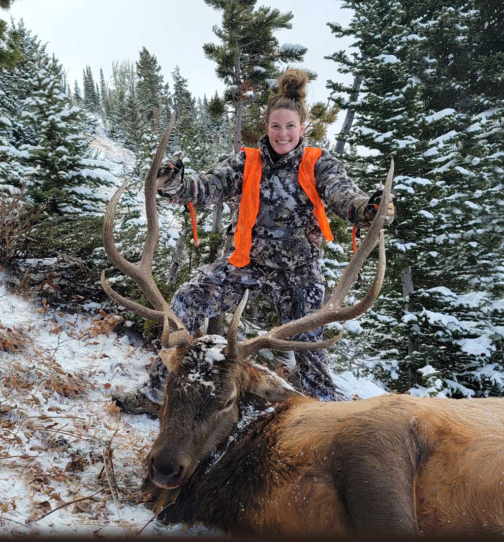 Woman in camouflage clothing and orange vest posing with dead elk after hunting