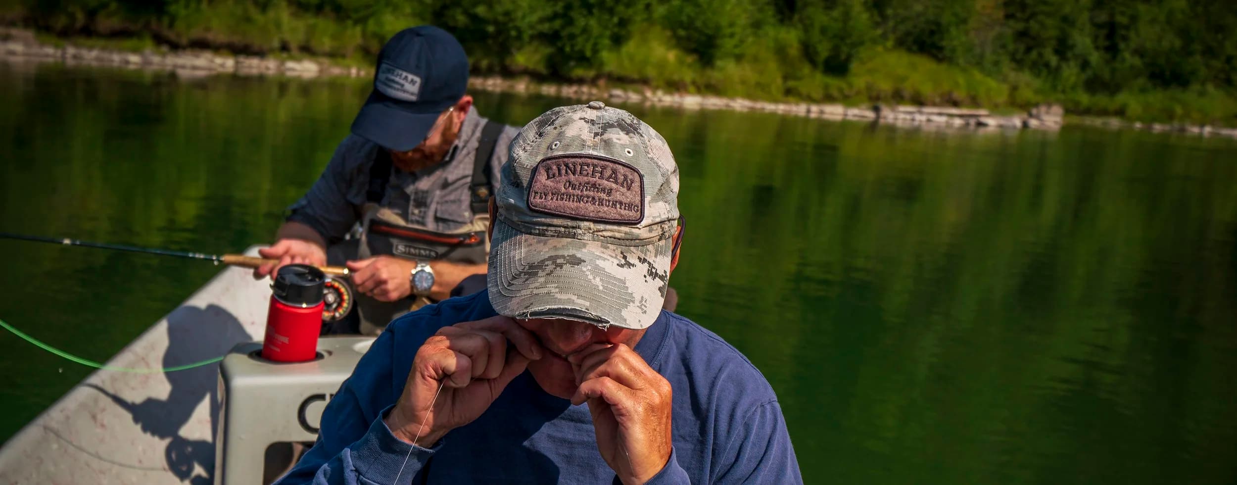 Two men fishing on a boat in a green lake with trees in the background, one wearing a camouflage cap that says "LINEHAN Outfishing Fly Fishing & Hunting" and the other in a navy cap.
