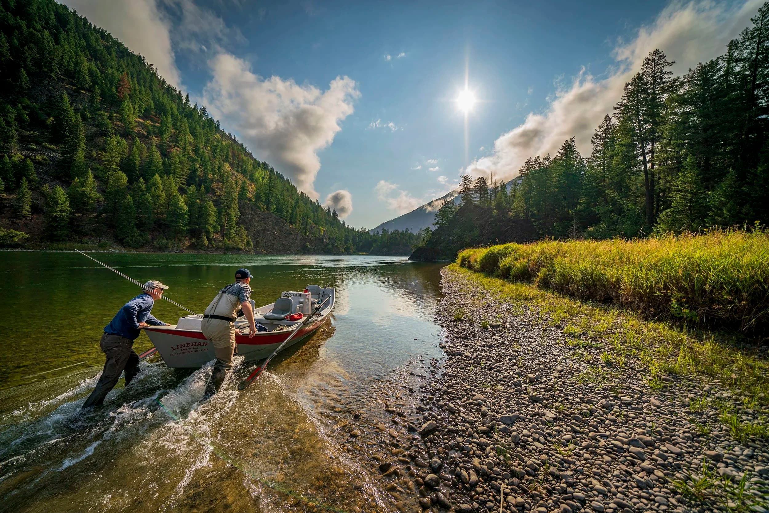 Two men launching a boat into a river in a mountainous and forested area under a bright sun with scattered clouds.